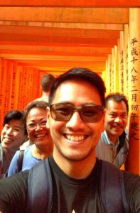 Jason's family is smiling beneath many bright red torii gates of Fushimi Inari Shrine. The gates are lined up one after the other in the background forming a tunnel.