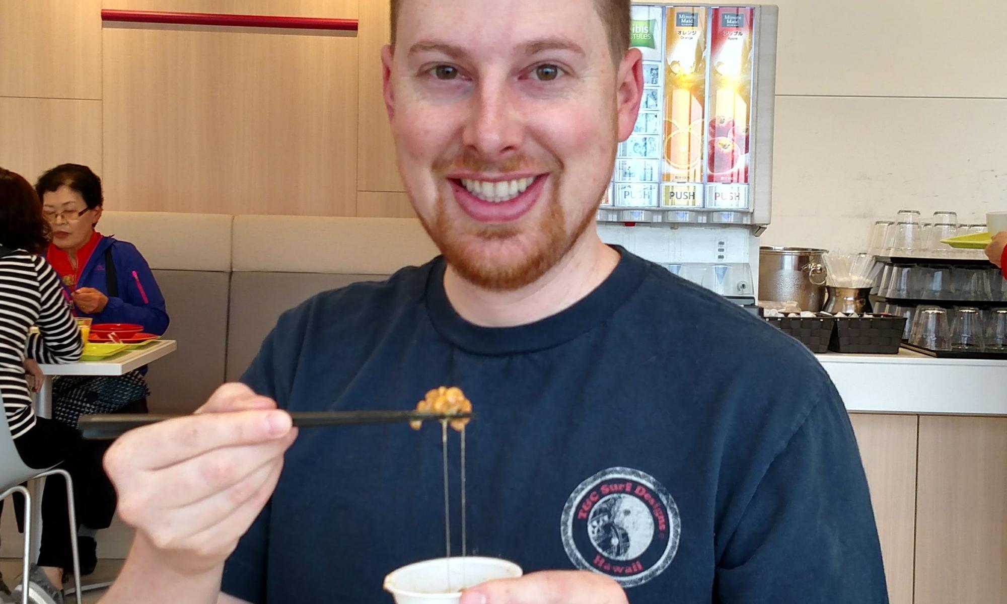 Matt is smiling with a small cup of natto in his left hand and chopsticks in his right hand. The soybean mush is gripped by the chopsticks with a trail of goo leading back into the cup. He is smiling despite the fact that he is about to eat something gross.