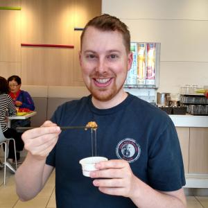 Matt is smiling with a small cup of natto in his left hand and chopsticks in his right hand. The soybean mush is gripped by the chopsticks with a trail of goo leading back into the cup. He is smiling despite the fact that he is about to eat something gross.