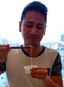Jason is not smiling while he holds up his cup of natto. His chopsticks also carry a lump of fermented soybeans with trailing goo leading back into the cup.