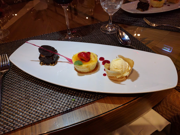 AmaWaterways Chef's Table dessert has three main pieces arranged on a rectangular white plate. On the left is a square chocolate truffle cake. The chocolate glazed exterior shimmers in the light. A thin, pink candy crystal leans across it. In the middle of the plate is a round brulée cheese cake. It is garnished with a mint leaf and strawberry with little dots of strawberry purée. On the right is a scoop of vanilla ice cream. It is sitting in a little wafer cup that tastes like an ice cream cone.